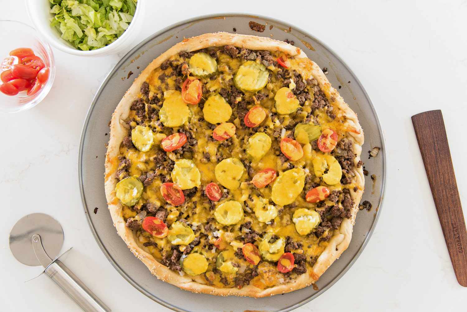 Baked Cheeseburger Pizza Next to a Bowl of Cherry Tomatoes and Another Bowl of Shredded Lettuce