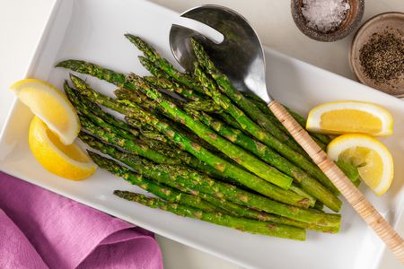air fryer asparagus on a platter with lemon slices at a table with a bowl of cracked pepper and a bowl of salt flakes