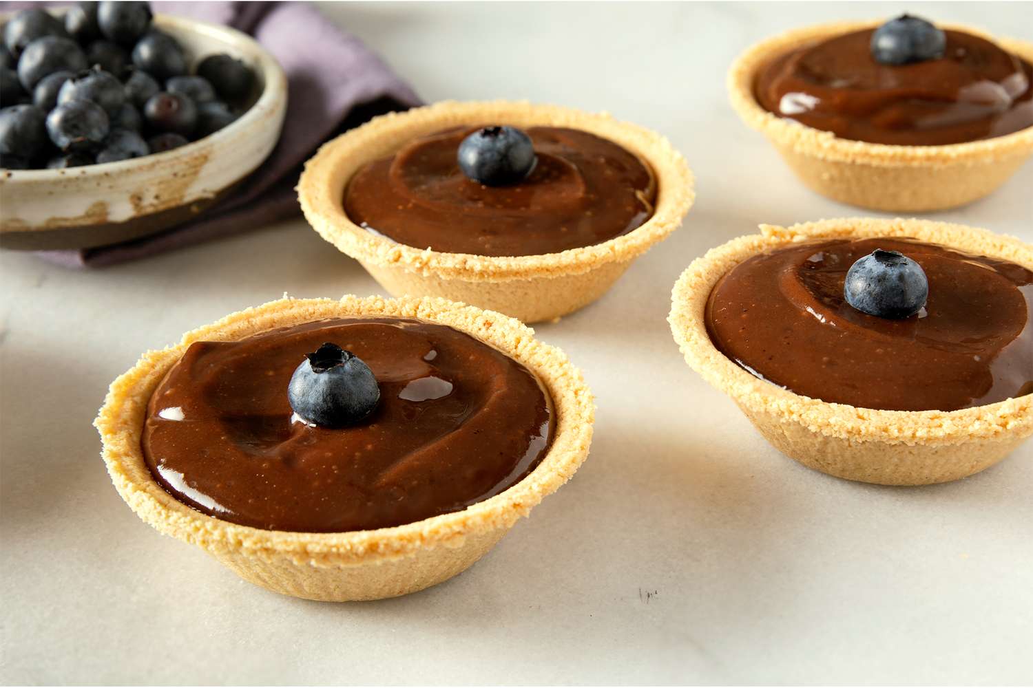 Mini chocolate pudding pies with a single blueberry on top set in graham cracker crusts arranged on a surface