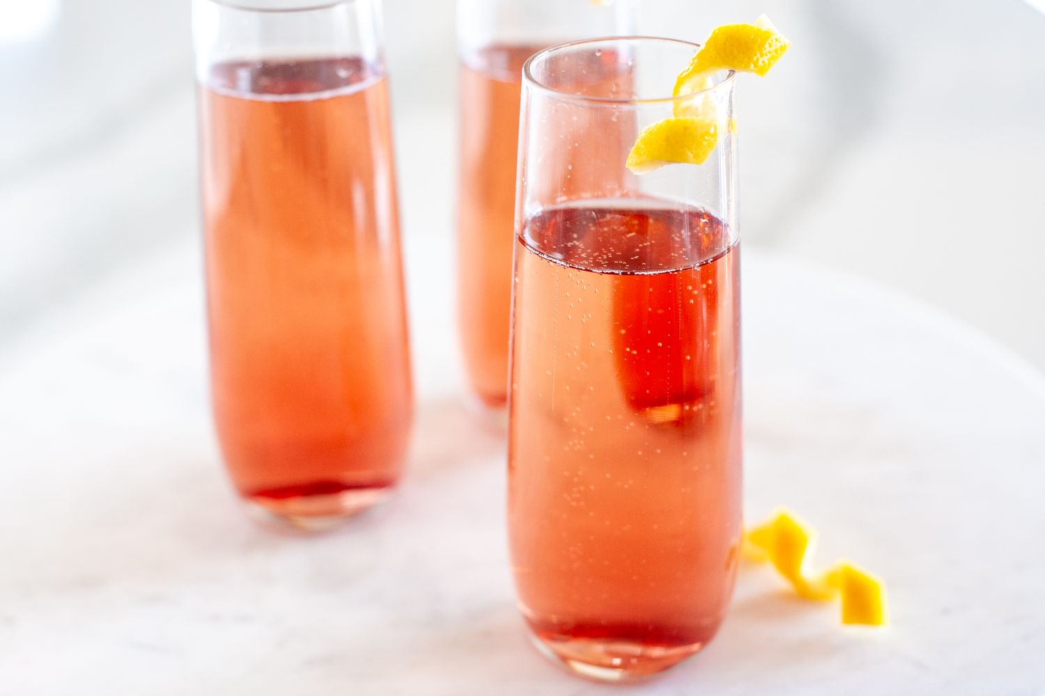 Three glasses of sparkling pink beverage garnished with orange peels served on a light marble surface