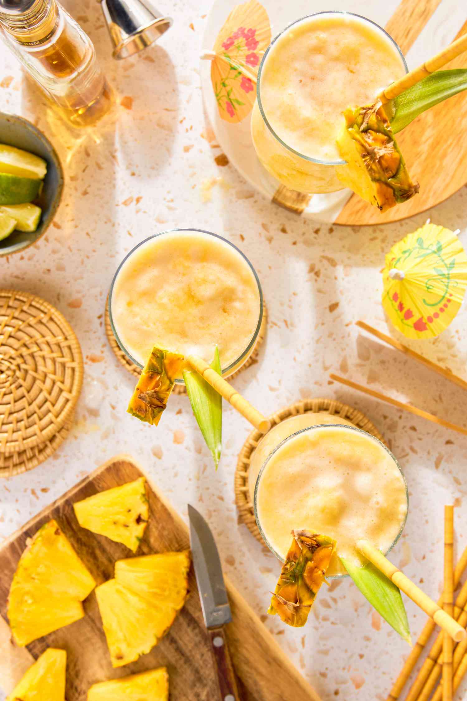 Overhead: Glasses of PiÃ±a Colada Served With a Pineapple Wedge, Pineapple Leaf, and a Straw (on Coasters), a Cutting Board With More Pineapple Wedges, a Bowl of Lime Edges, Straws and a Jigger on the Counter, and a Bottle of Rum