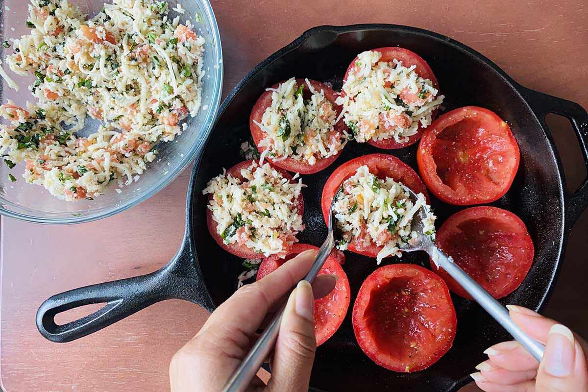 Halved and seeded tomatoes set in a cast iron skillet and being filled to show how to make stuffed tomatoes.
