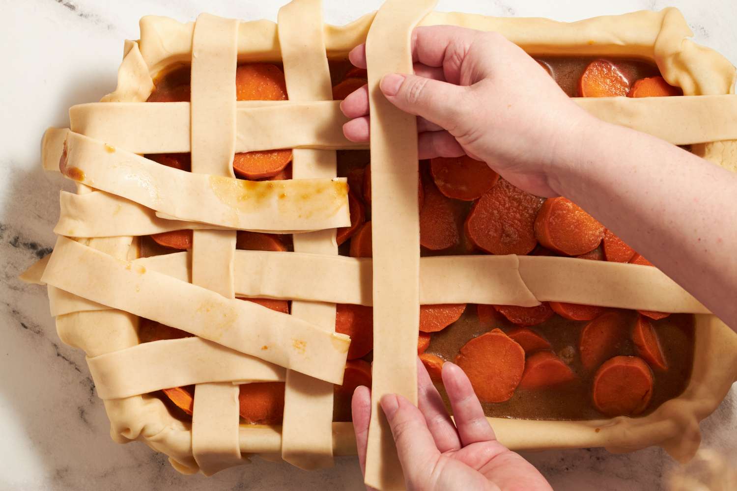 Hands arranging lattice strips on top of a dish filled with sliced sweet potatoes in the process of creating the sweet potato sonker recipe