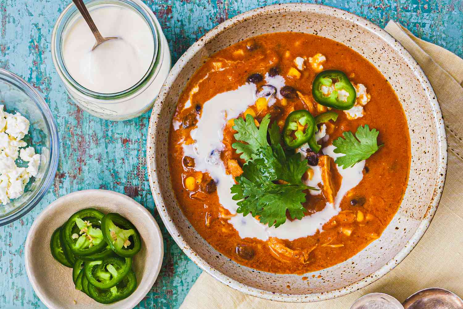 Bowl of Creamy Chicken Enchilada Soup Garnished with Cilantro, Crema, and Jalapenos Surrounded by Little Bowls with Toppings (Jalapenos, Lime Wedges, Cotija, and Crema)