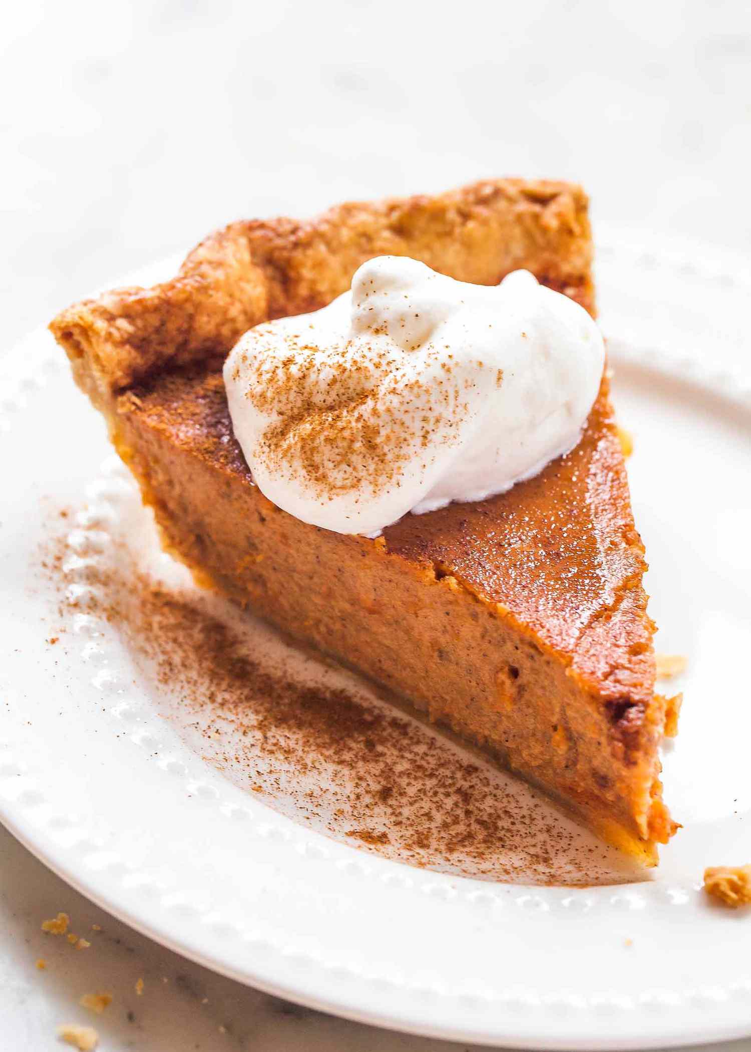 Slice of sweet potato pie on a white plate with whipped cream dusted with cinnamon and fork.