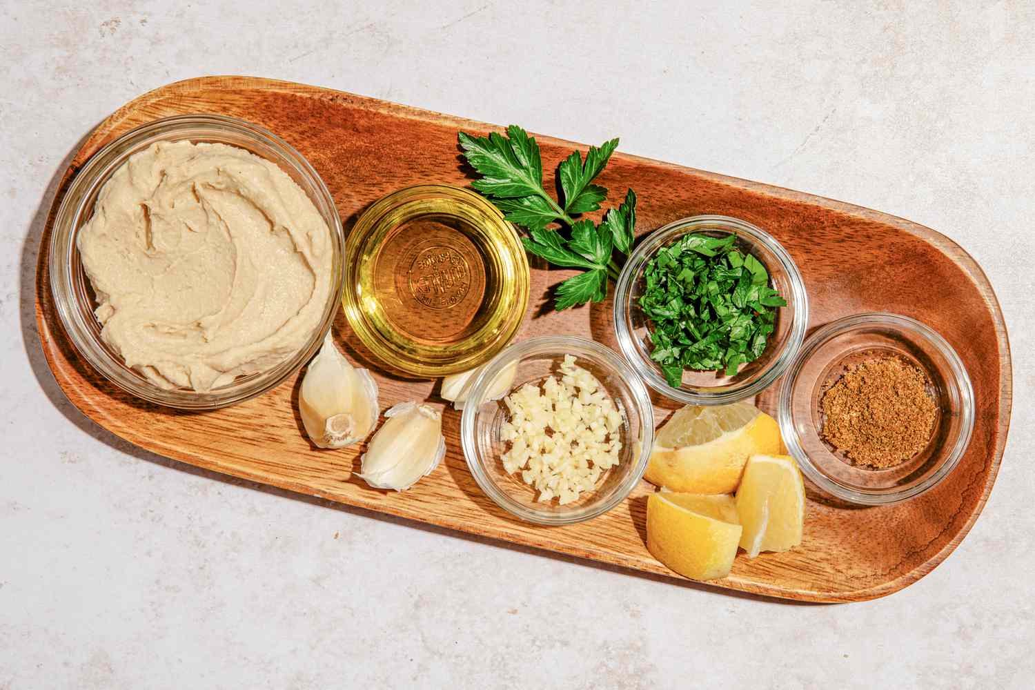 Bowls of hummus salad dressing ingredients (hummus, olive oil, minced garlic, minced parsley, ground cumin - each in it's own bowl), cloves of garlic, lemon wedges, and a sprig of parsley, all on a wooden tray