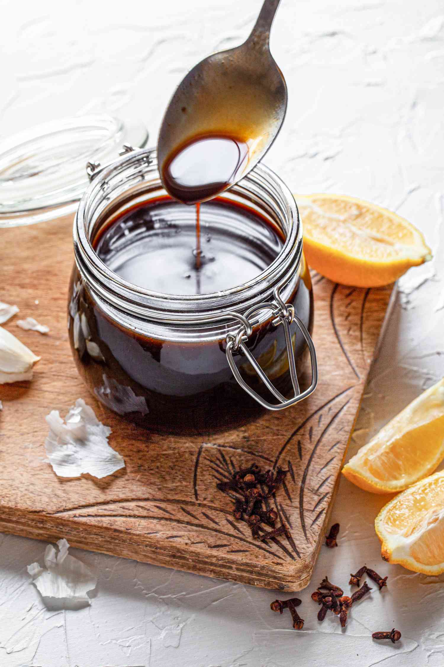 Spoonful of Worcestershire Sauce Lifted from a Jar Sitting on a Decorative Wooden Board Covered in Ingredients (Halved Orange, Coriander Seeds, Garlic Cloves, Cloves, and Lemon Wedges)