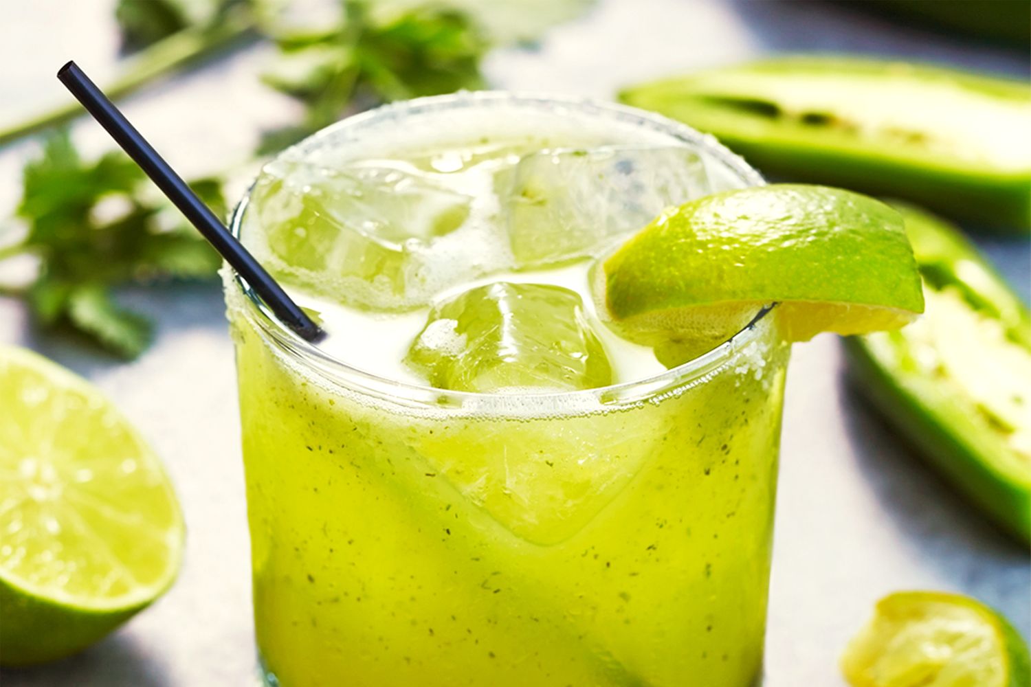 Glass of a green margarita with garnish, surrounded by lime slices and herbs