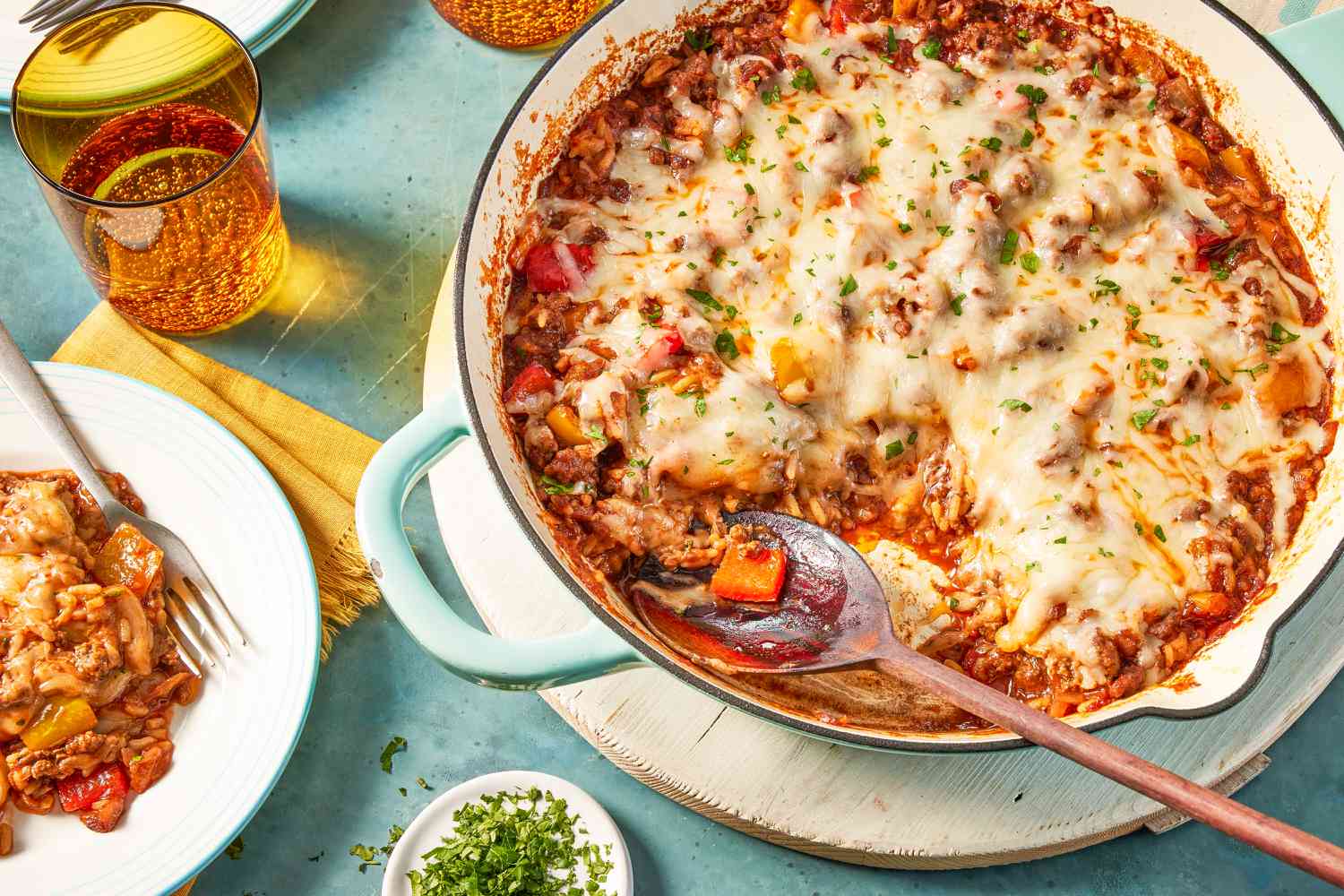 unstuffed peppers in a skillet at a table setting with a serving on a plate, a glass of water, a small dish with minced herbs, and a stack of plates