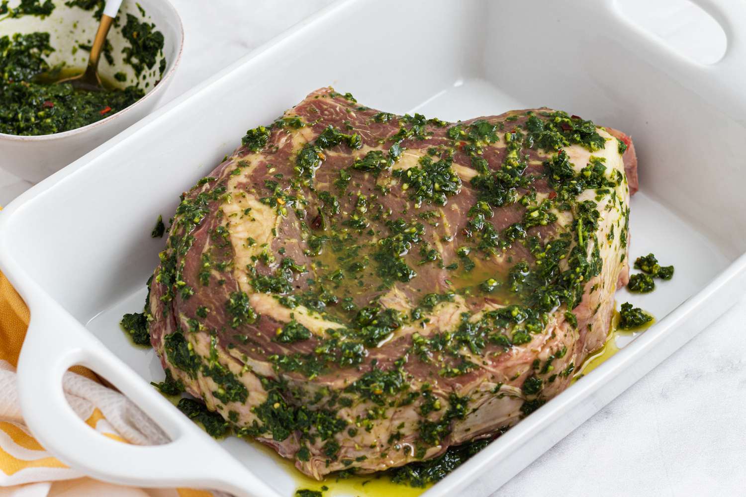 Chimichurri sauce spread on top of a cowboy steak in a baking dish.