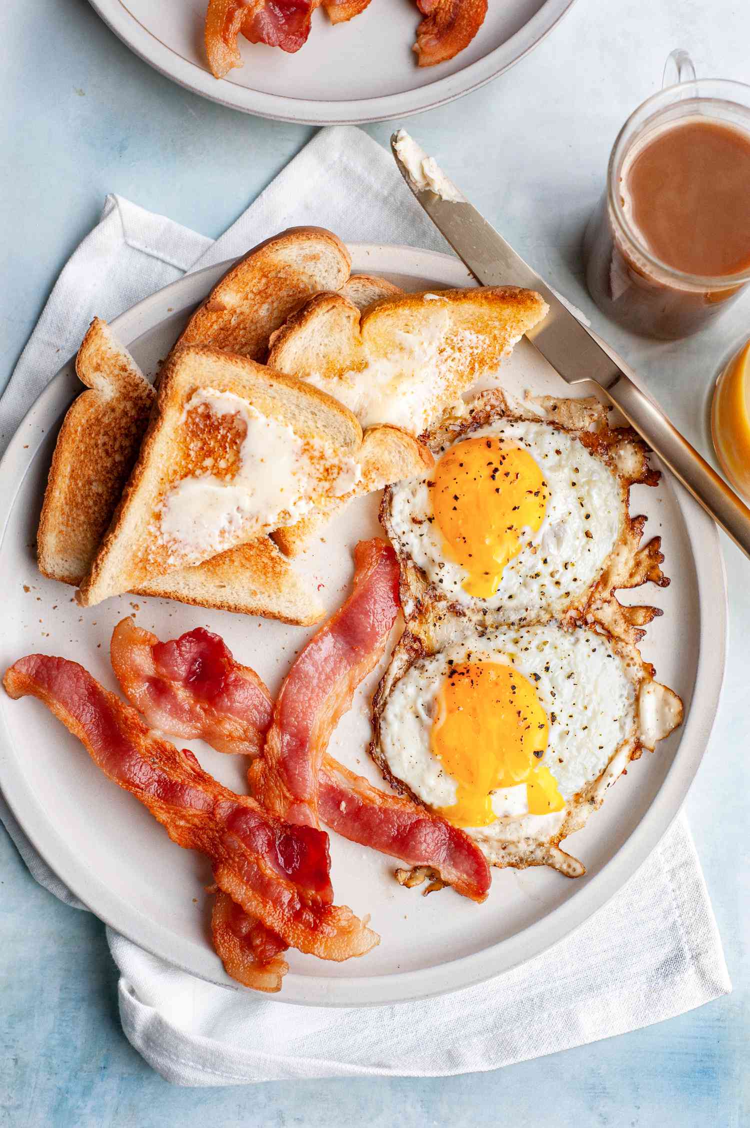 Plate of Microwave Bacon with Sunny Side Eggs and Slices of Buttered Toast, Surrounded by a Glass of Orange Juice, Mug of Coffee, and a Plate of Bacon
