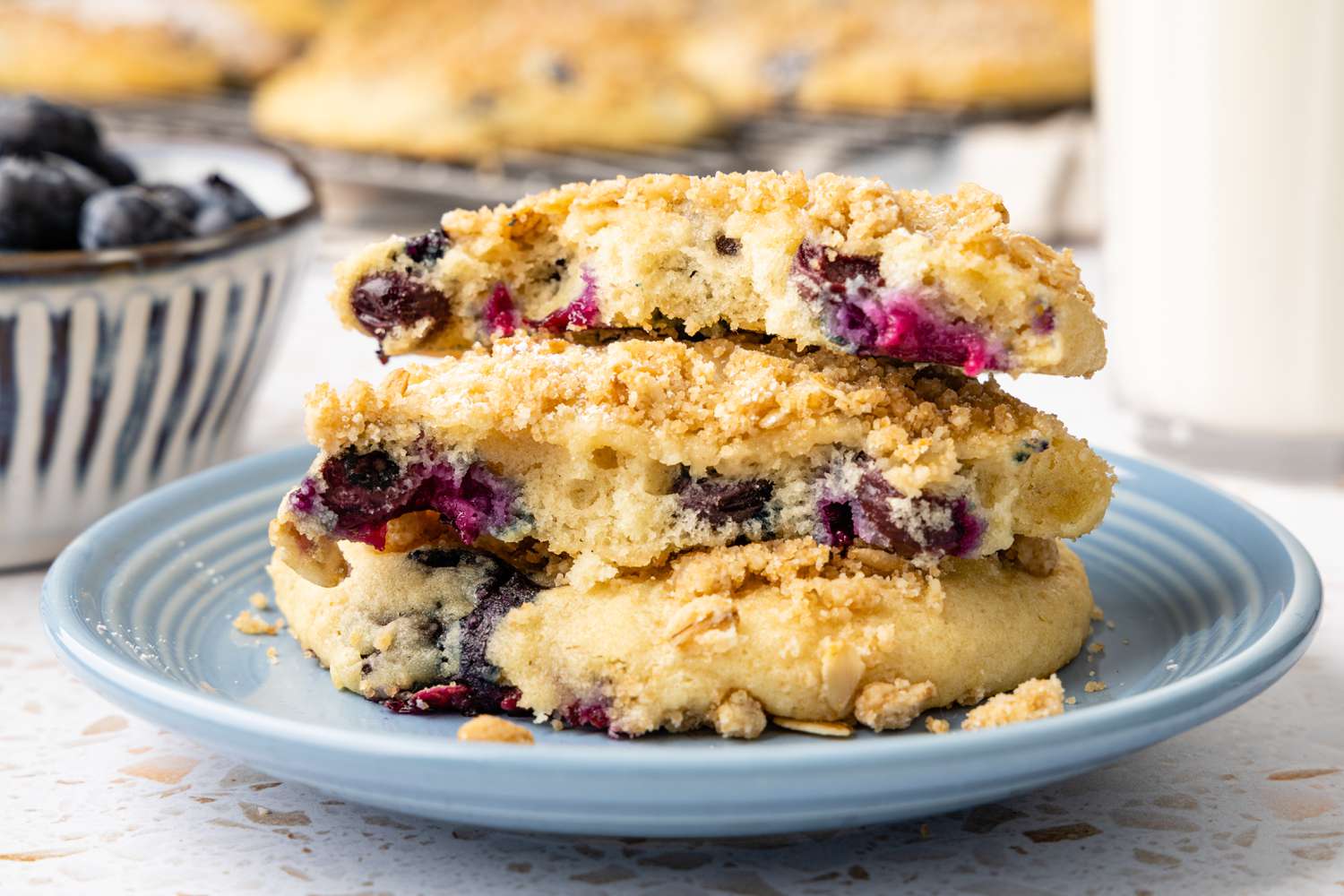 Stack of Blueberry Muffin Tops on a Plate with the Top Two Muffin Tops Broken in Half, and in the Background, a Bowl of Blueberries, More Muffin Tops on a Wire Rack, and a Glass of 