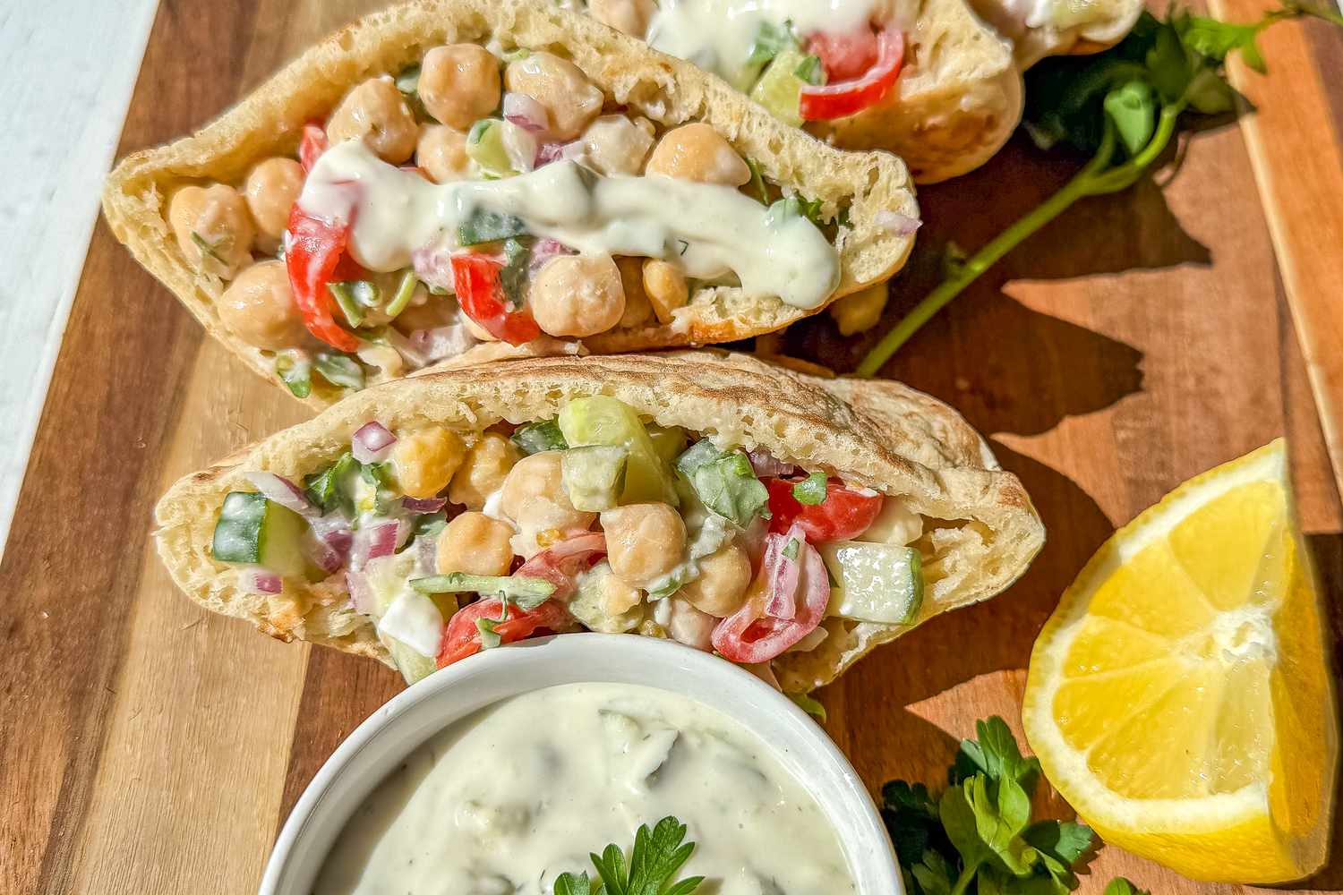 Greek salad pockets next to a some olive, fresh herbs, a halved lemon, and small bowl of tzatziki, all on a cutting board 
