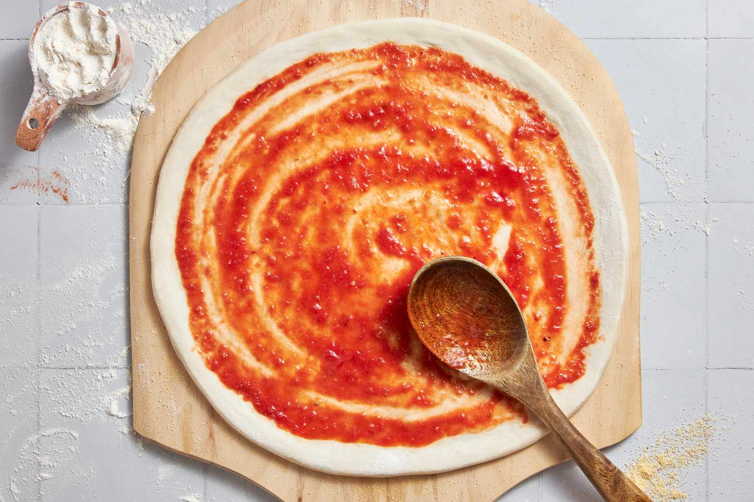 Sauce Added to Shaped Pizza Dough on a Wooden Pizza Peel, and on the Counter Next to It, a Cup of Flour for Vegan Pizza Recipe