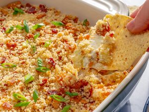 Closeup of a baking dish with jalapeño popper dip, and a hand holding a tortilla chip scooping some of the dip out