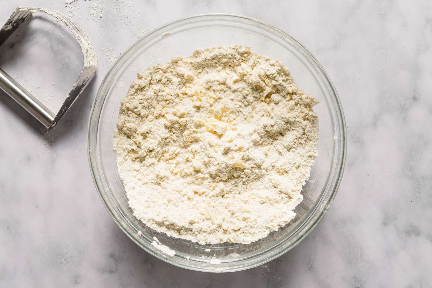 A glass bowl containing a flour and butter mixture on a marble countertop with a pastry cutter nearby for Chocolate Chip Scones recipe