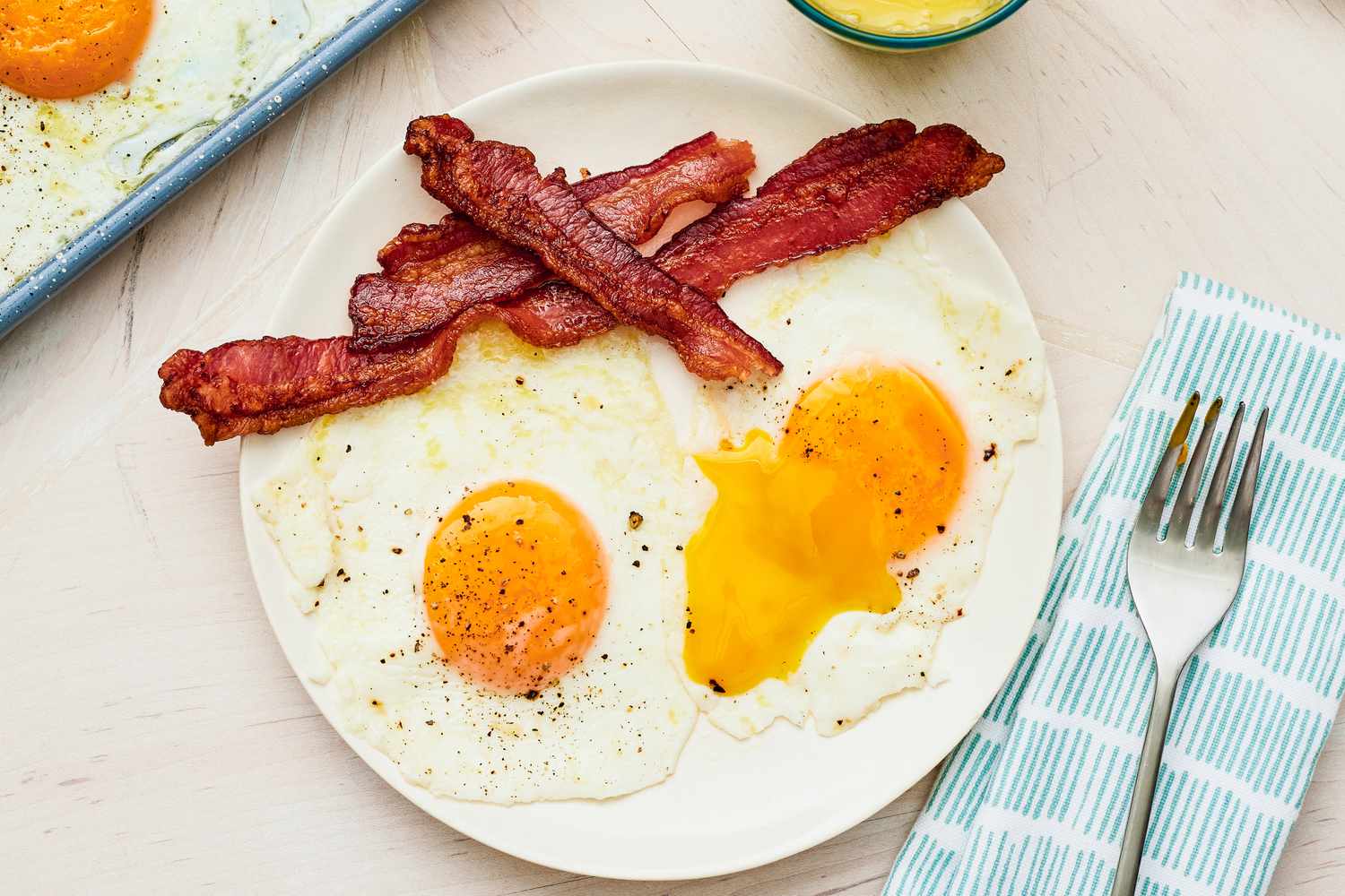 Plate of Sheet Pan Fried Eggs Served With Bacon, and in the Surroundings, a Glass of Orange Juice, a Fork on a Table Napkin, and More Sheet Pan Fried Eggs on a Sheet Pan