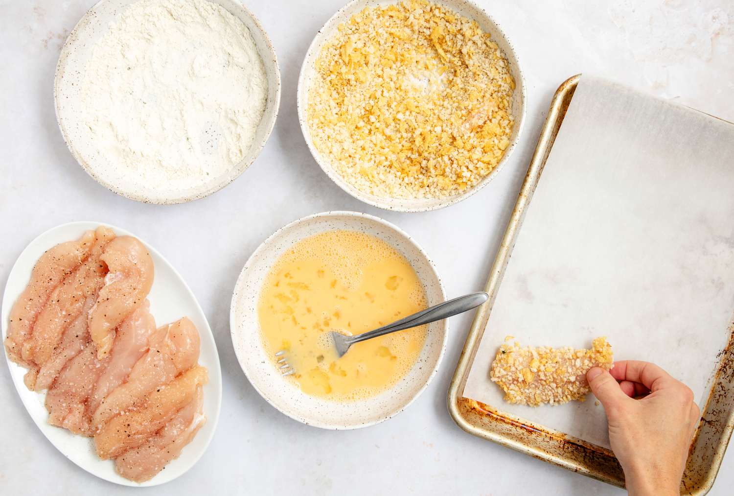 hand putting battered chicken tender on a small parchment lined baking pan; bowl of egg mixture, breadcrumb mixture, and flour, and a plate of raw seasoned chicken tenders