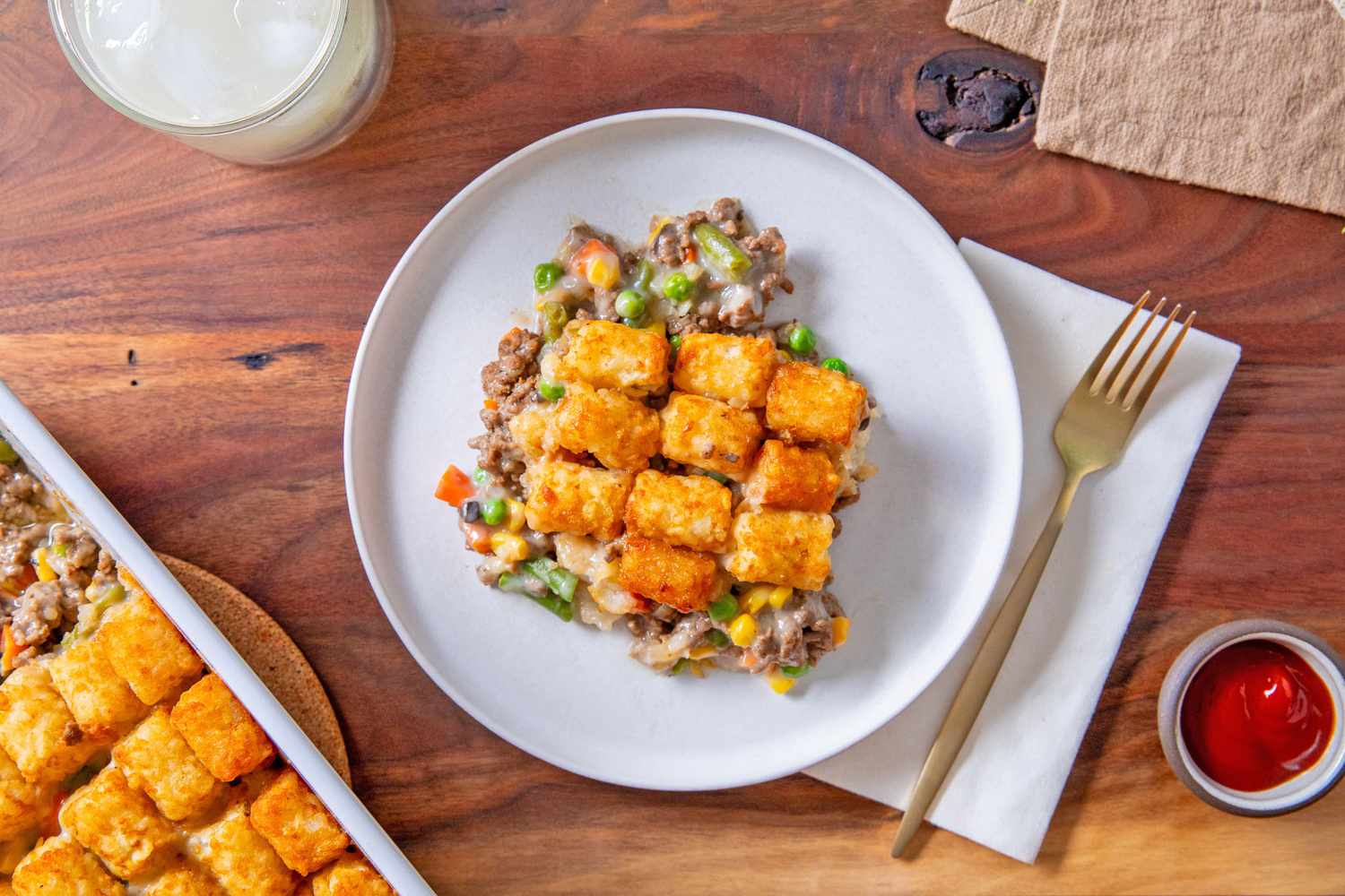 A serving of Classic Tater Tot Hotdish on a plate with a fork and napkin and a small dish of ketchup