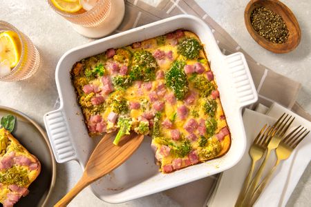 Overhead view of a white casserole dish of broccoli cheese pie with a serving spoon in a table setting with forks, glasses and a bowl of pepper to the side