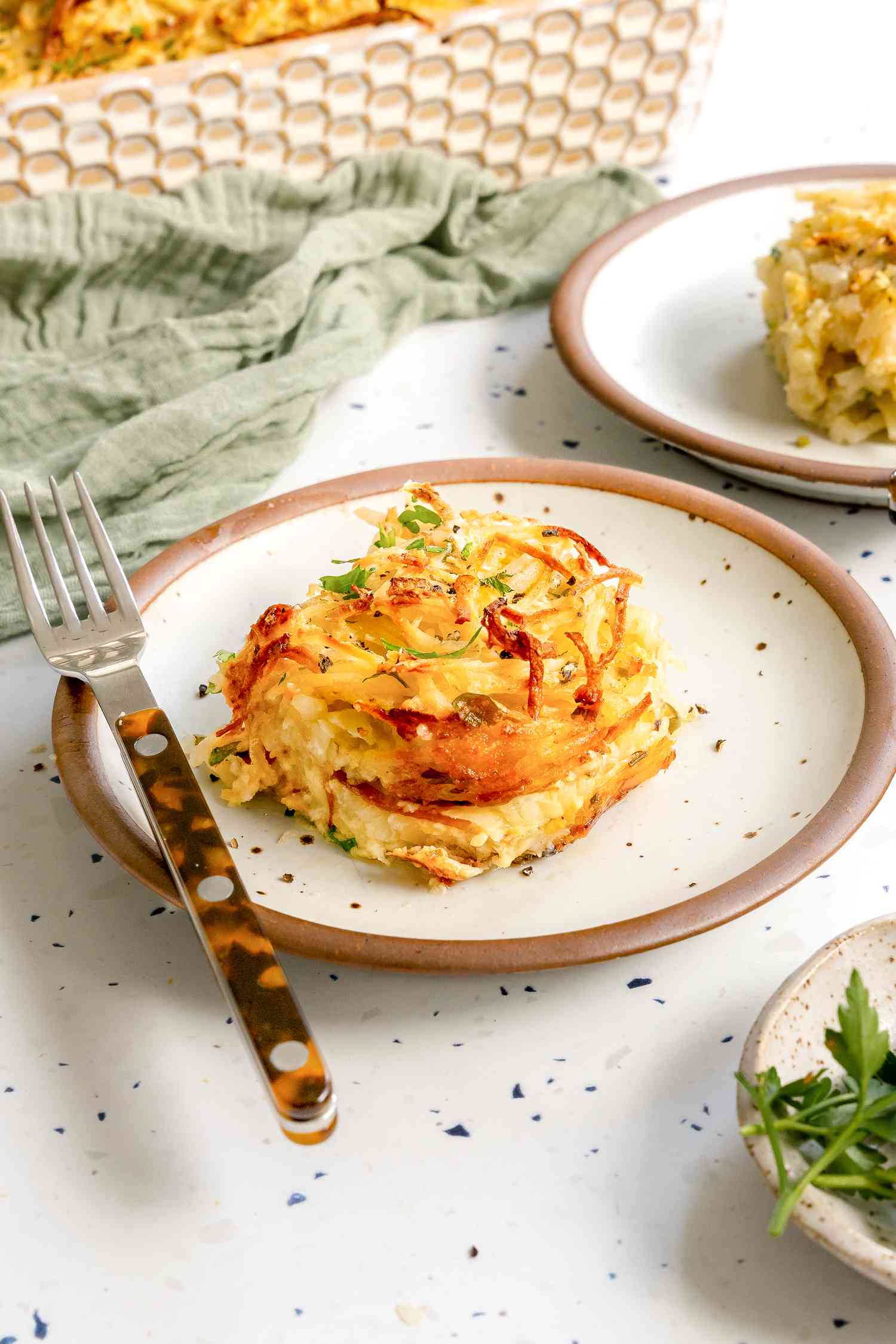 Serving of Potato Kugel on a Plate with a Fork, and Surrounding It, Another Plate with Another Serving, a Small Saucer with Herbs, a Kitchen Linen, and the Casserole Dish with More