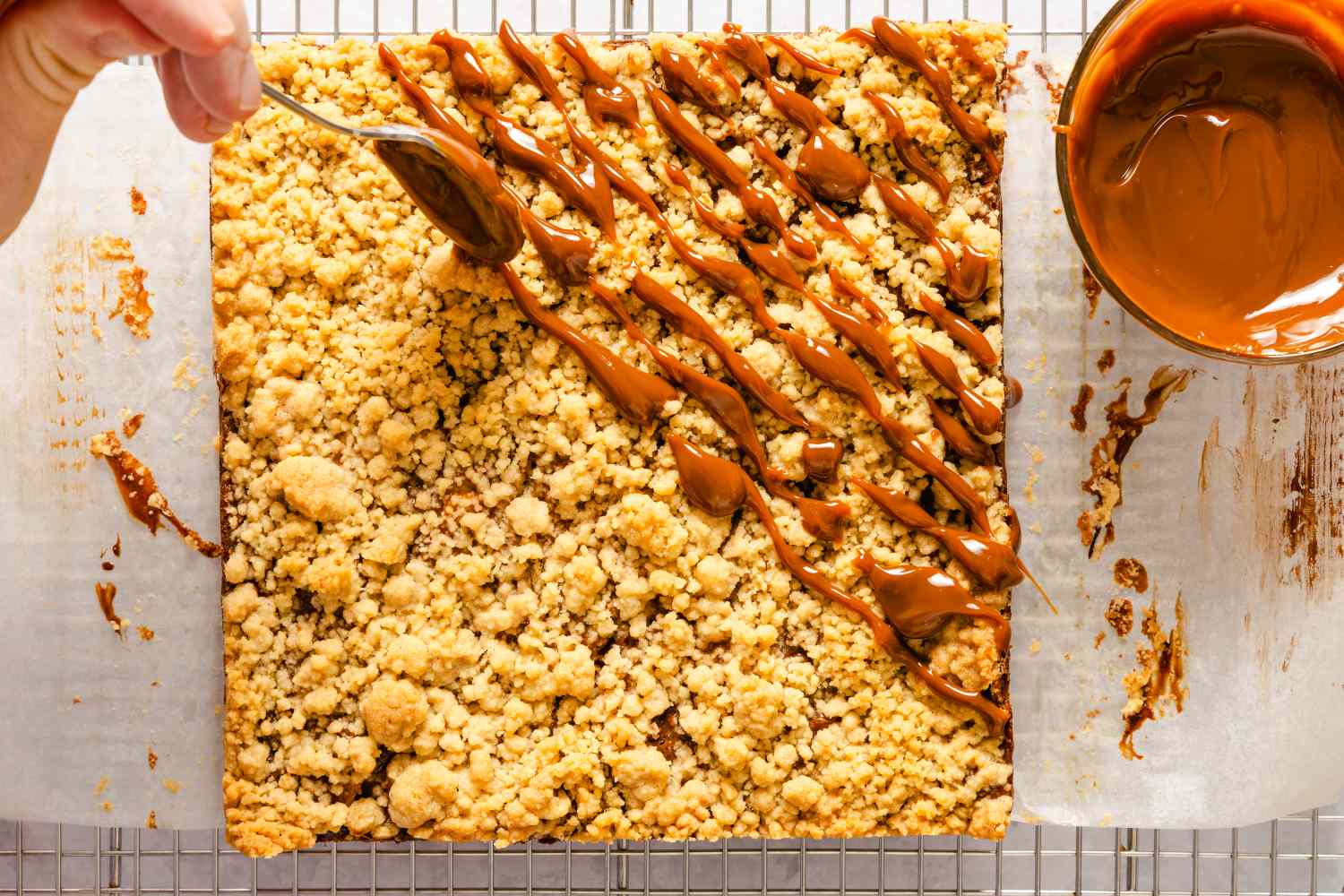 Hand drizzling caramel sauce from a spoon onto caramel apple bars recipe on parchment paper