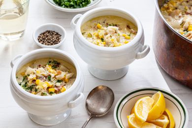 Corn and Crab Chowder in Two Soup Bowls with Handles at a Table Setting with a Pot with More Chowder, a Glass of White Wine, a Plate of Parsley, Small Bowl of Cracked Pepper, and Bowl of Lemon Slices