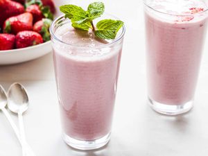 Two tall glasses of healthy strawberry almond oat smoothies on a white counter. A sprig of mint is on the top of one glass two spoons and a bowl of strawberries are to the left of the glasses.