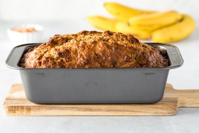 Loaf of banana bread in a baking pan on a wooden board with bananas in the background