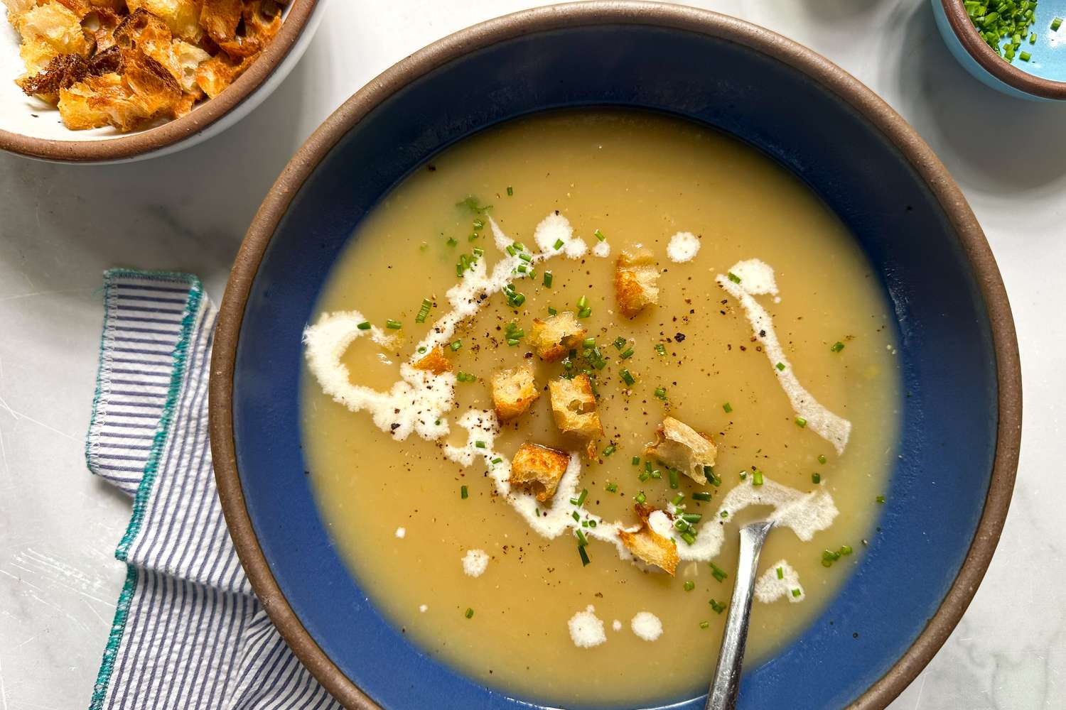 A shallow bowl of potato leek soup topped with a drizzle of cream, croutons, and green herbs