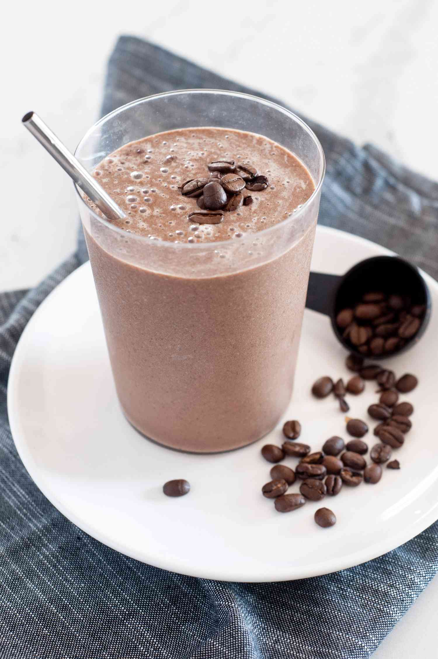 Creamy mocha coffee smoothie with a straw and set on a white plate with coffee beans next to it.