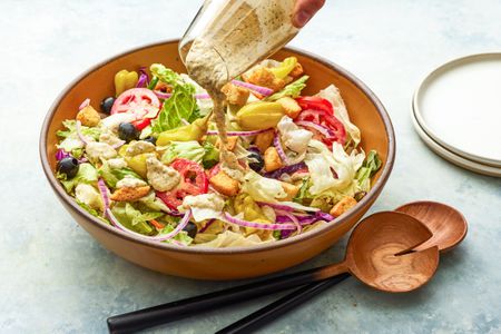 Salad Dressing Poured Over a Bowl of Olive Garden Salad, and on the Counter Next to It, a Stack of Plates and a Set of Salad Serving Utensils