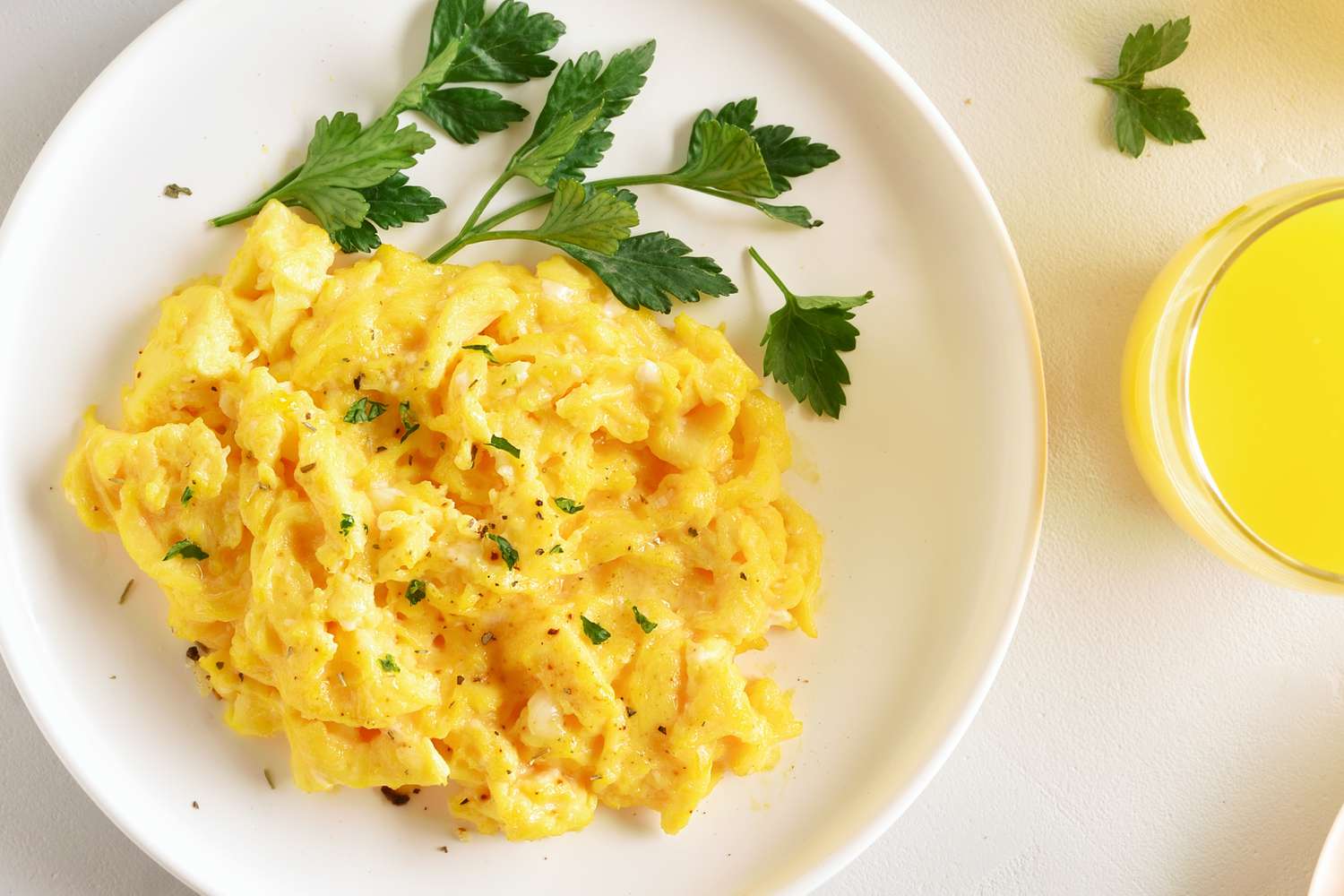 Golden scrambled eggs on a plate with a sprig of parsley, next to a small glass of orange juice
