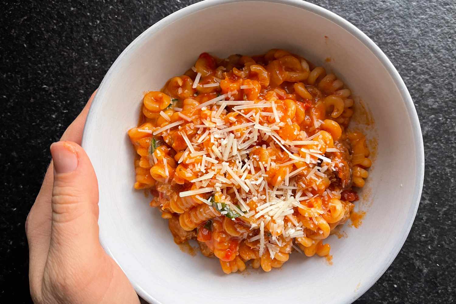 A person holding a bowl of rotini pasta with tomato sauce and grated cheese
