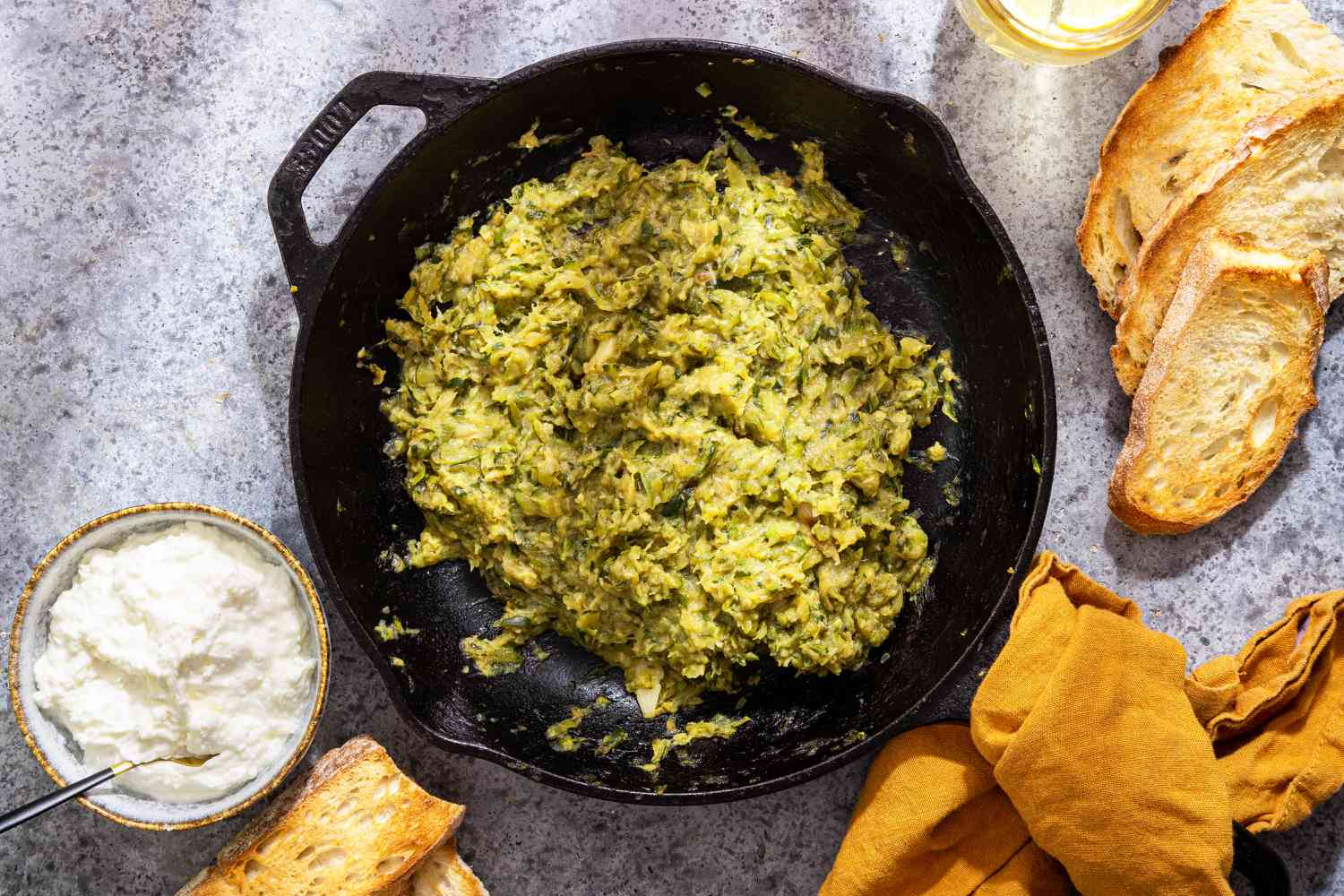 Zucchini Butter in a Cast Iron Skillet With a Wooden Spoon, and in the Surroundings, a Mustard Colored Kitchen Towel, Pieces of Toast, Fresh Thyme Sprigs, a Glass of Water, and a Bowl of Ricotta