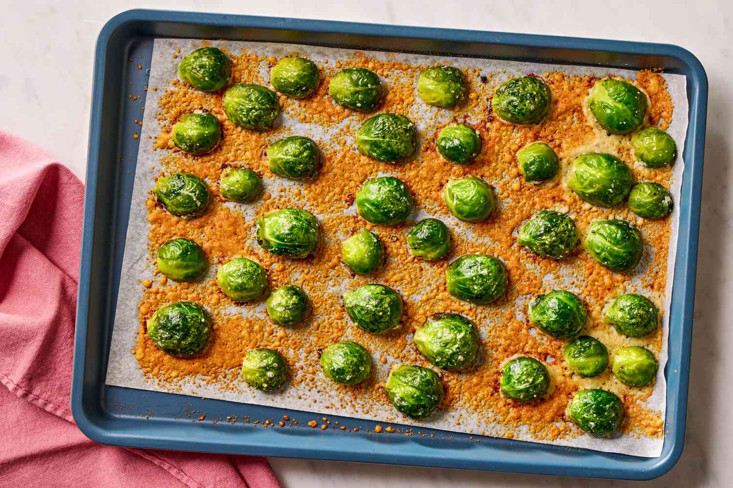 Overhead view of a blue baking sheet lined with parchment paper with cooked brussels sprout slices and parmesan for Crispy Parmesan Brussels Sprouts Recipe