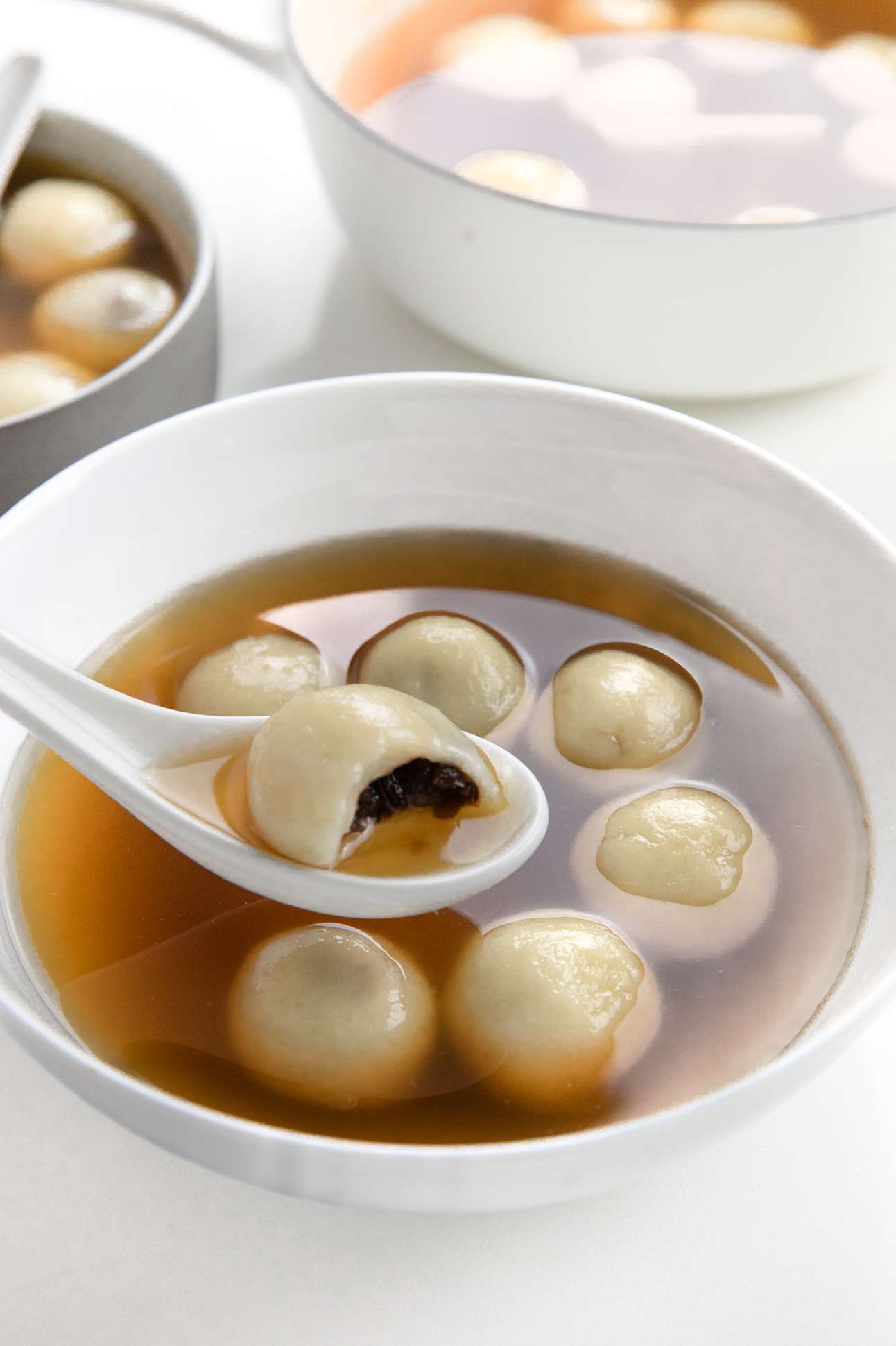 Bowl of Red Bean Tangyuan with One Tangyuan Dumpling Sitting on a Raised Soup Spoon with a Bite Missing, and in the Background, Another Bowl and More in a Pot 