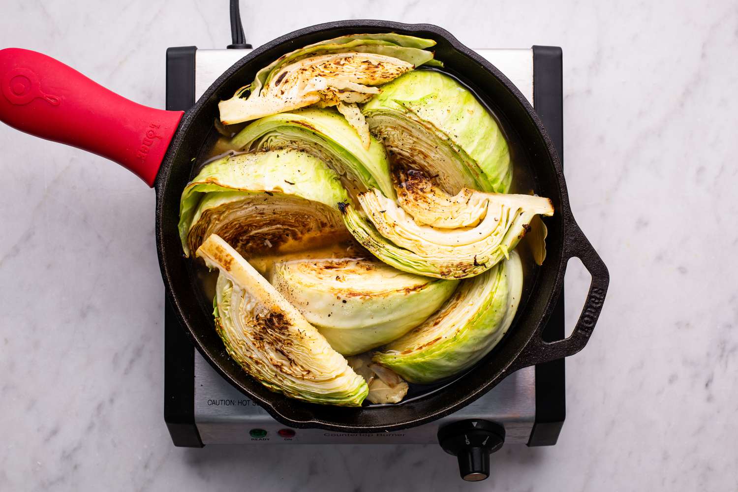 Overhead view of a cast iron skillet cooking cabbage wedges for Melting Cabbage recipe on a hot plate on a marble countertop