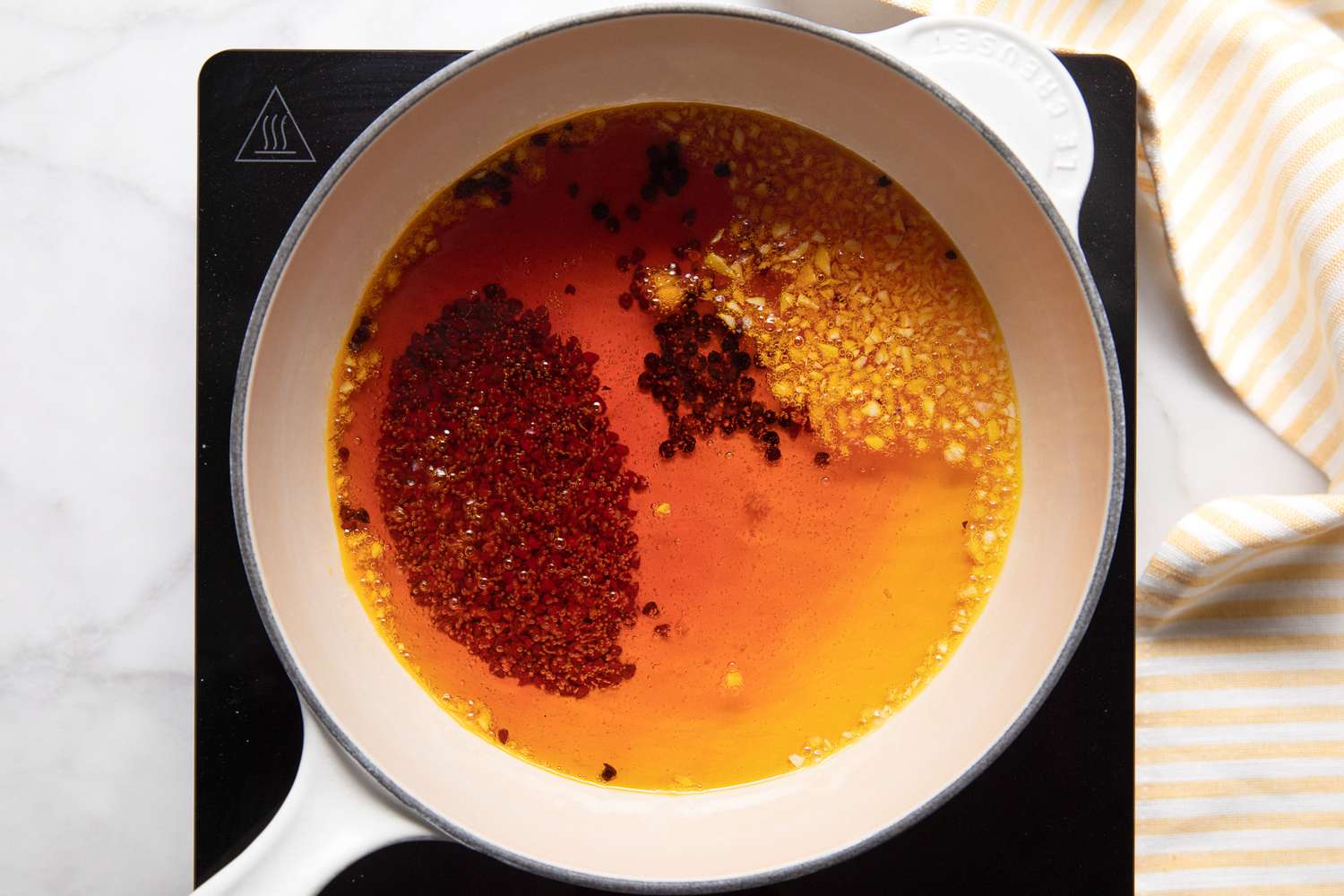 Annatto Oil Ingredients Steeped in a Pot Over a Portable Burner Heated Until Color Is Almost "Blood Orange", and on the Counter Next to It, a Yellow and White Striped Kitchen Towel for Banana Ketchup Recipe
