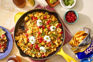 Frito pie dip in a cast iron skillet at a table setting with a bag of fritos lays, a single serving of dip on a plate, a small bowl of diced tomatoes, and a beer