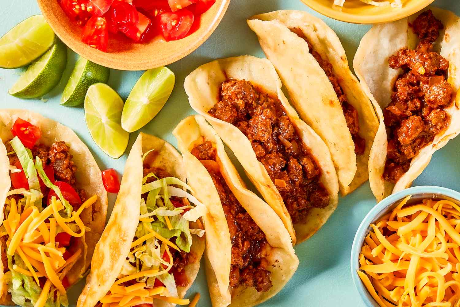 An assortment of tacos with beef shredded lettuce shredded cheese garnishes and lime wedges