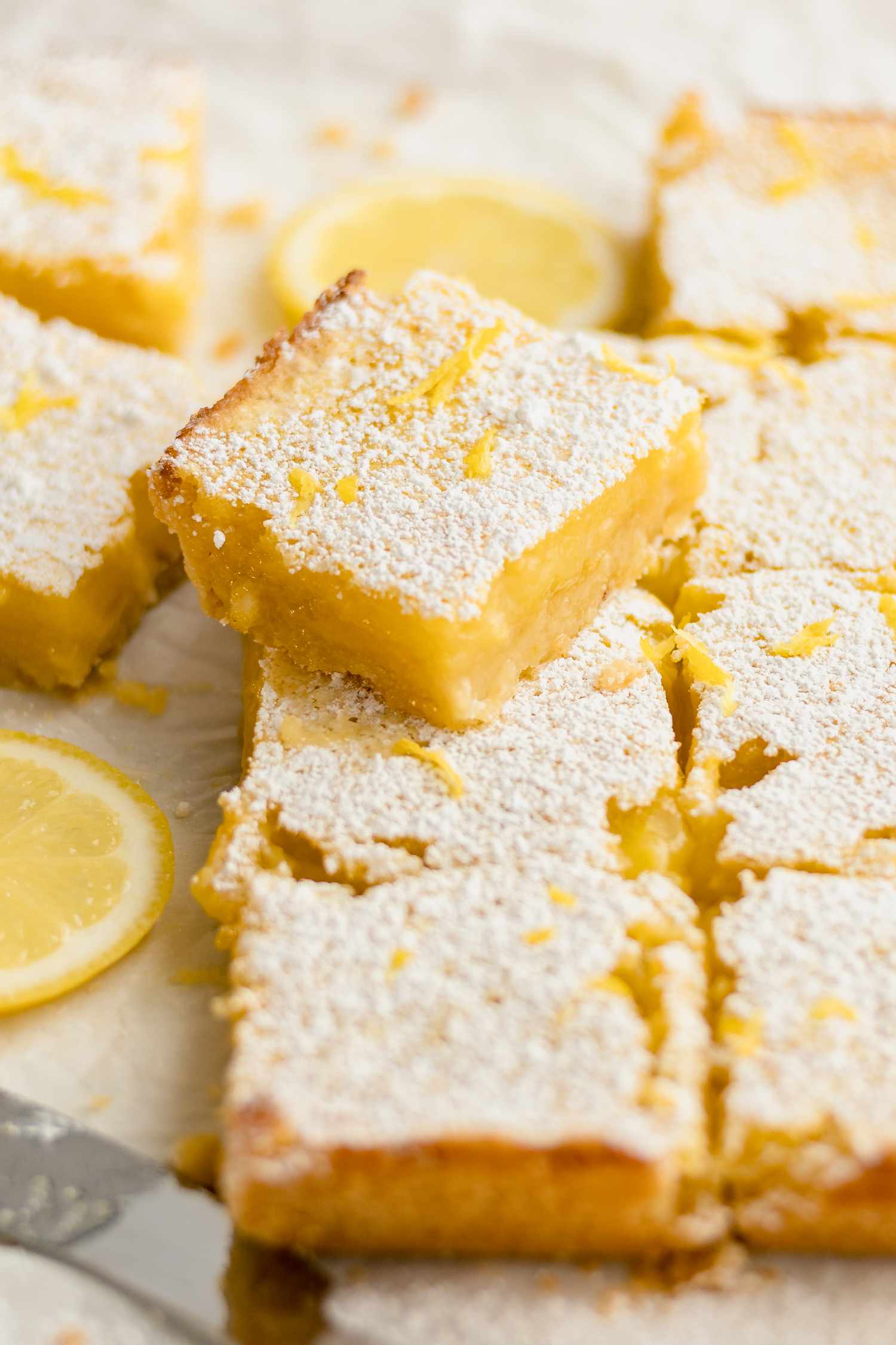 Lemon Bars Covered with Lemon Zest. Some Are Spread Out on a Parchment Surface with One Stacked Next a Lemon Slice.