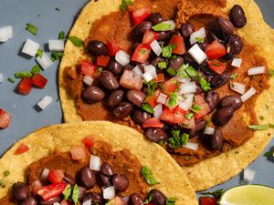 Two tostadas topped with refried beans black beans diced tomatoes onions and herbs on a plate with a lime wedge nearby