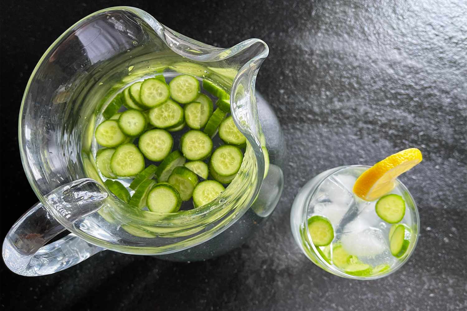 A pitcher of water with cucumber slices and a glass of infused water with a lemon garnish on a dark surface