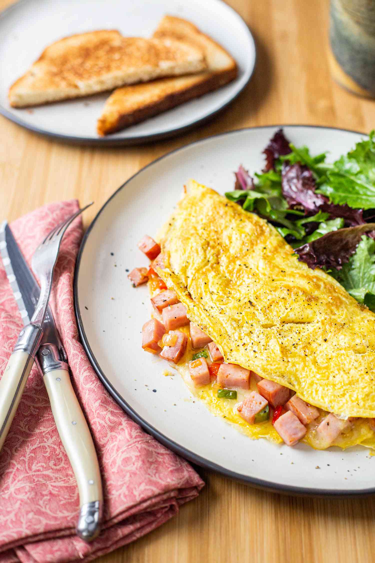 Denver Omelet with Mixed Greens on Plate Next to Utensils on Napkin and Plate of Toast in Background