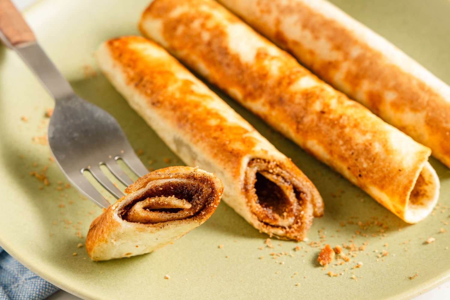Three rolled cinnamon sugar tortillas on a plate with one cut in half, revealing the filling along a fork