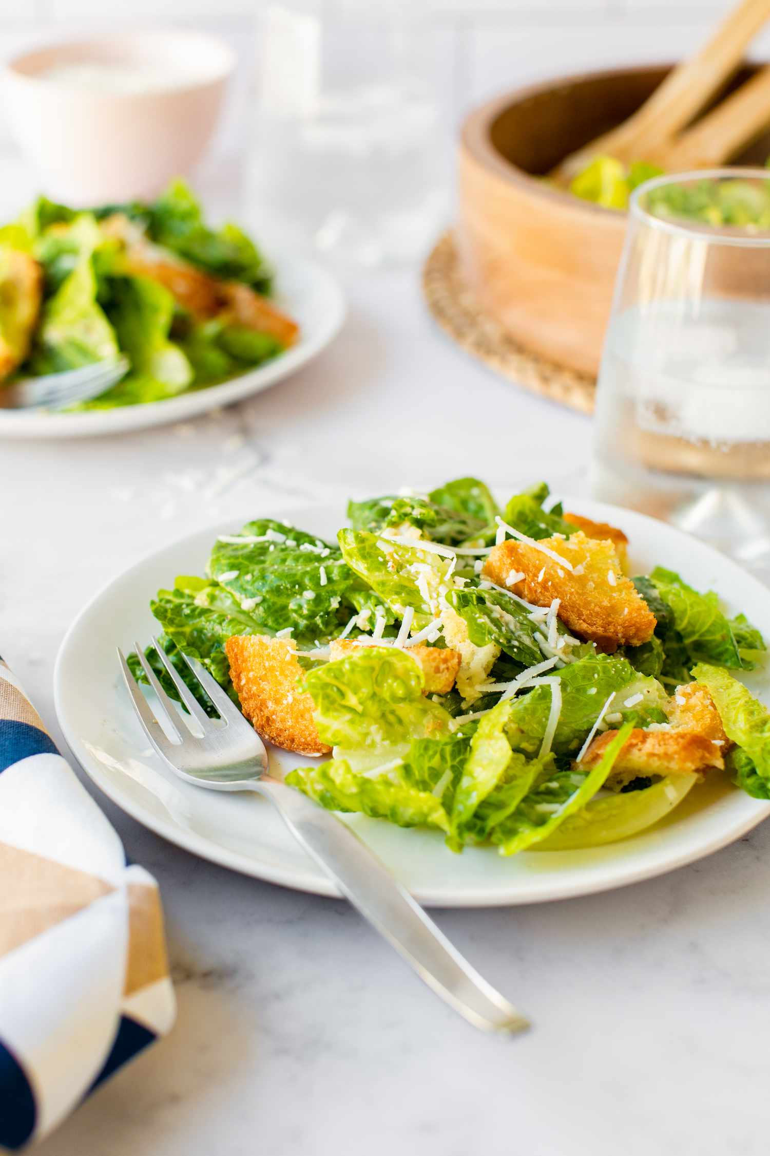 A Dining Table Spread Consisting of Multiple Plates with Classic Caesar Salad, Bowl of More Salad, a Glass of Water, and a Table Napkin