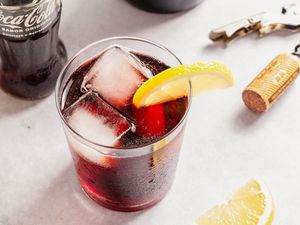 A glass of Kalimotxo with ice cubes and a lemon wedge, a cola bottle and a wine cork on the side