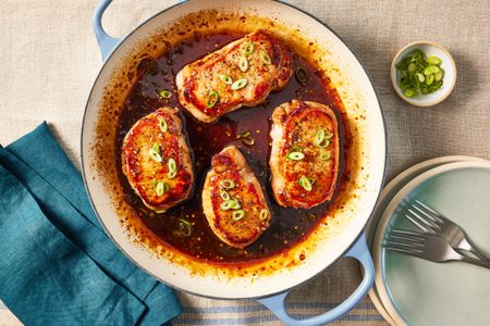 glazed pork chops in a wide skillet at a table setting with a stack of bowls, a table napkin, and a small bowl of sliced scallions