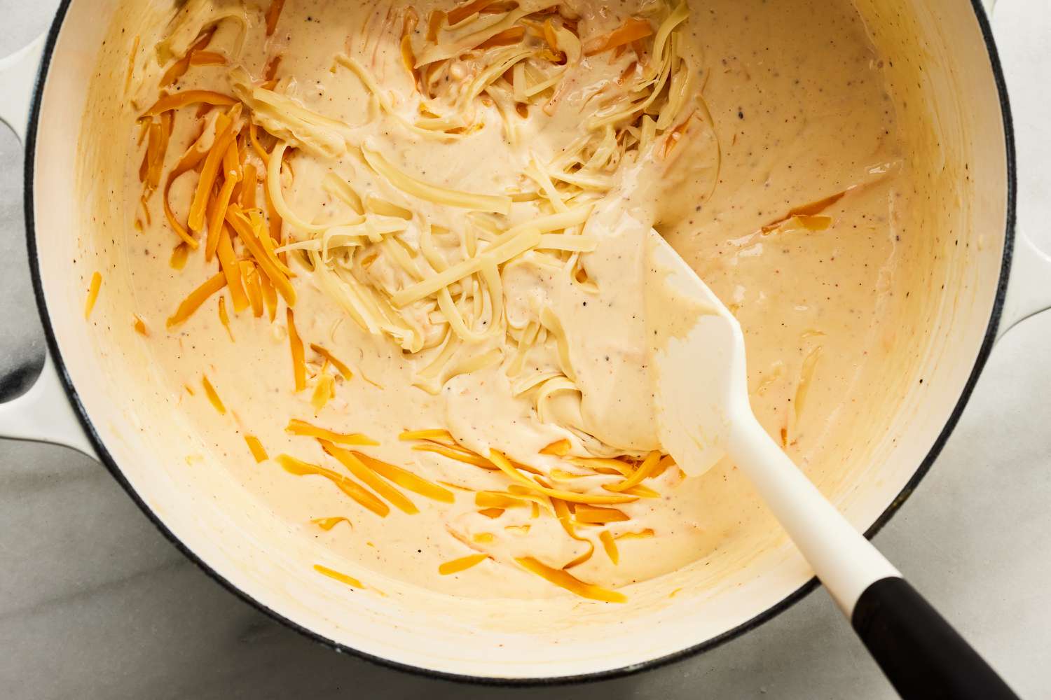Overhead view of a white dutch oven while mixing the cheese into the cream sauce with a spatula for Million Dollar Mac and Cheese recipe